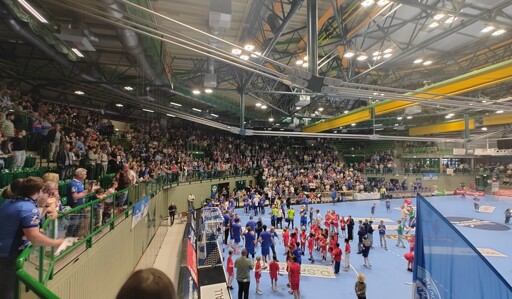 Das Bild zeigt eine große, gut besuchte Handballhalle. Im Vordergrund befindet sich ein blauer Spielfeld, auf dem Spieler in blauen und roten Trikots versammelt sind. Die Spieler scheinen sich zu versammeln, möglicherweise nach einem Spiel. Im Hintergrund sind zahlreiche Zuschauer auf den Tribünen zu sehen, die das Spiel verfolgen. Die Halle ist gut beleuchtet und hat eine helle, offene Struktur mit einem hohen Gewölbe und einem offenen Dach. Die Tribünen sind mit grünen Sitzreihen ausgestattet, und die Zuschauer sind engagiert und begeistert. Auf der rechten Seite des Bildes ist ein großes blaues Banner zu sehen, dass das Logo des Vereins BHC zeigt. Die Atmosphäre wirkt lebendig und voller Energie.